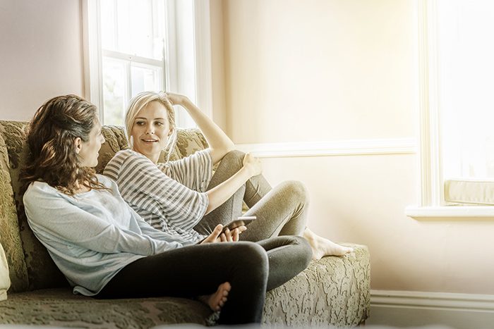 A photo of female friends gossiping on sofa. Happy young women looking at each other. Both are in casuals at home. They are in brightly lit room.