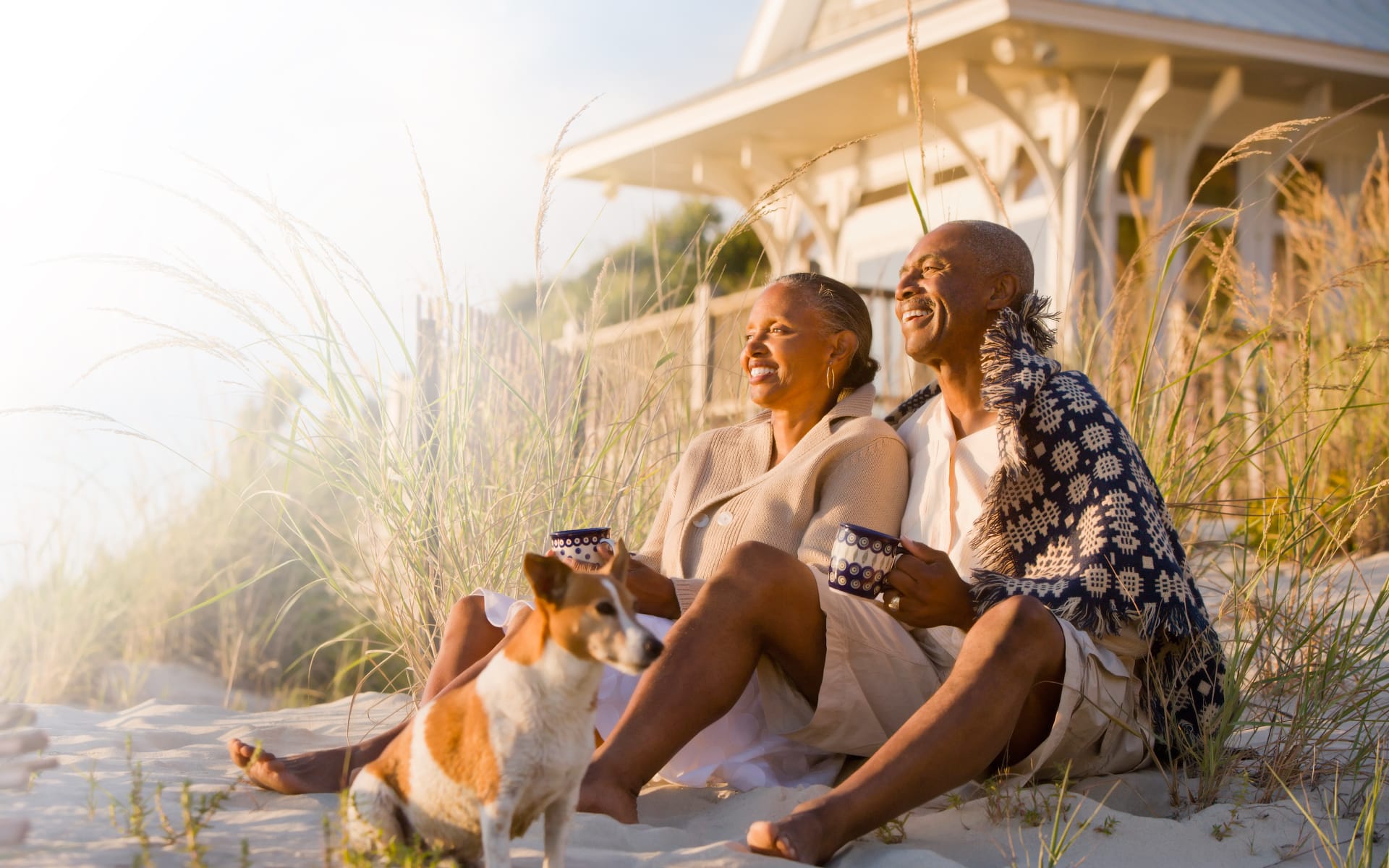 Couple sitting on beach smiling with dog drinking coffee and watching the ocean sunrise.