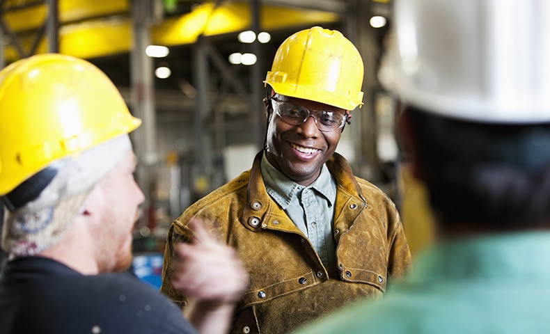 Three industrial workers in hard hats are conversing on the storeroom floor. The African-American man faces the camera, which caught the backs of the other two. He is wearing a brown jacket over a blue denim shirt. The man on the left has on a navy shirt. The one to the right has on a teal shirt.