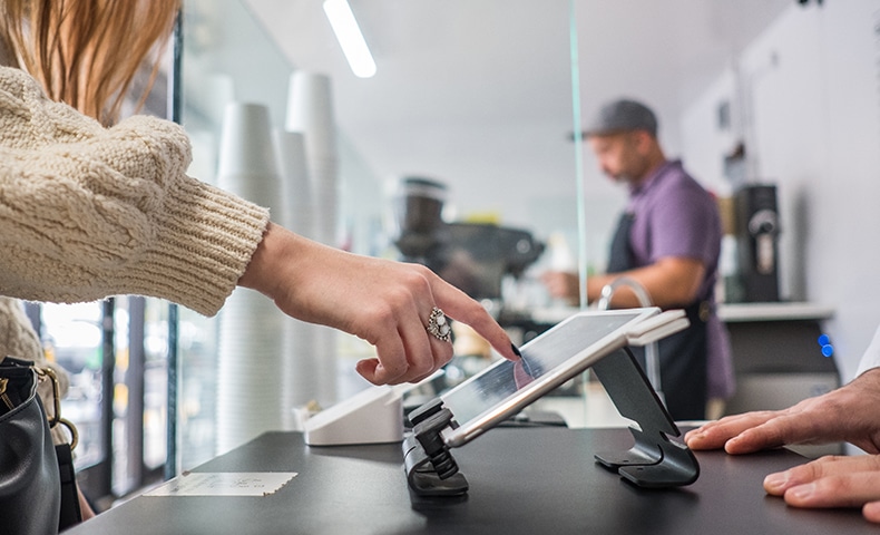 Profile view of a young woman signing the bill on the digital tablet at the register at a local neighborhood coffee shop.