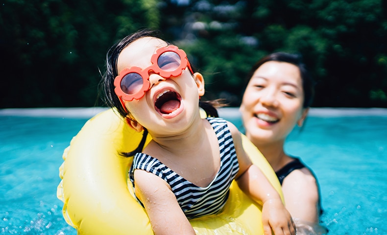 Happy Asian toddler girl with sunglasses smiling joyfully and enjoying family bonding time with mother having fun in the swimming pool in summer