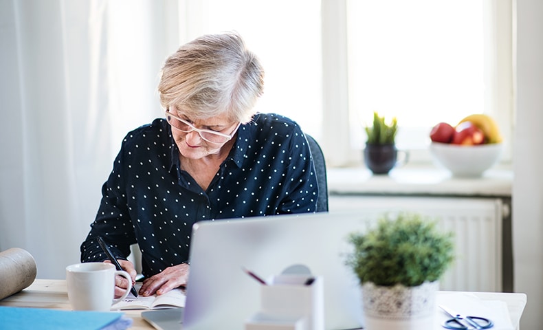 An active senior woman with laptop working in home office, writing.